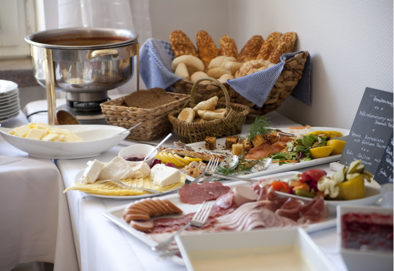 Frühstücksbuffet im Hotel mit Brötchenkorb und Porzellanplatten mit unverpackten Lebensmitteln wie Butter, Käse und Wurst-Aufschnitt