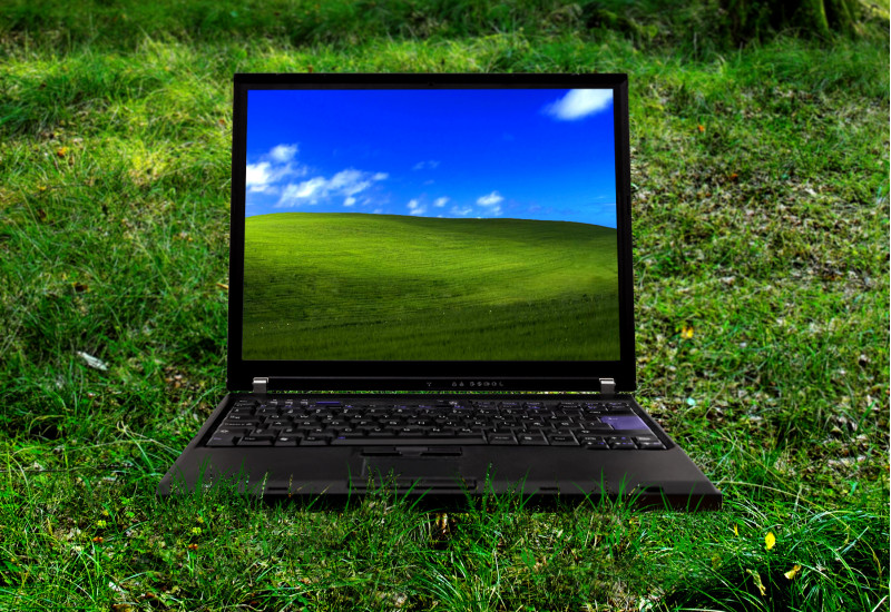 ein Laptop steht auf einer grünen Wiese, auf dem Bildschirm ist ein grüner, grasbewachsener Hügel vor blauem Himmel zu sehen