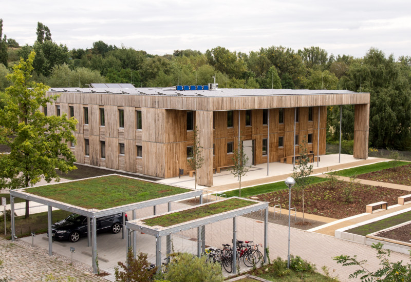 quadratischer, zweistöckiger Flachbau mit Holzfassade und Fahrradständerüberdachungen mit Dachbegrünung inmitten frisch angelegter und bepflanzter Außenanlagen