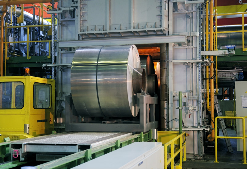 in einer Fabrikhalle wird ein große Rolle aufgerolltes Aluminiumband auf einem Förderband in eine Kammer gefahren, in der es glüht