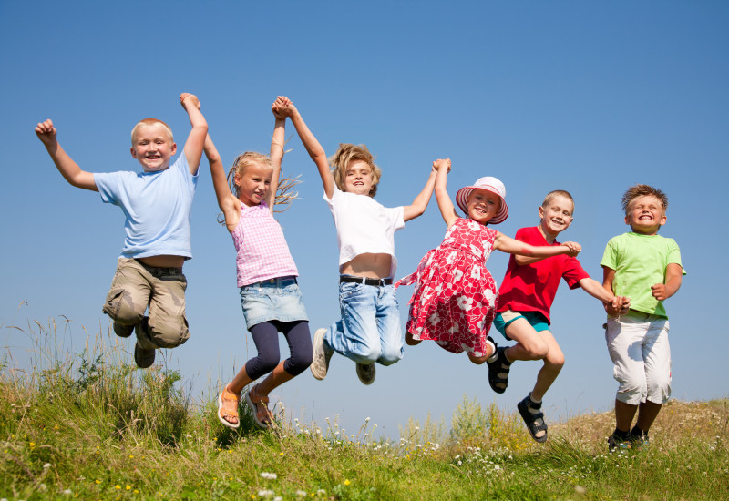 Sechs Kinder auf einer blühenden Wiese vor blauem Himmel haben sich in einer Kette bei den Händen gefasst und hüpfen lachend in die Luft