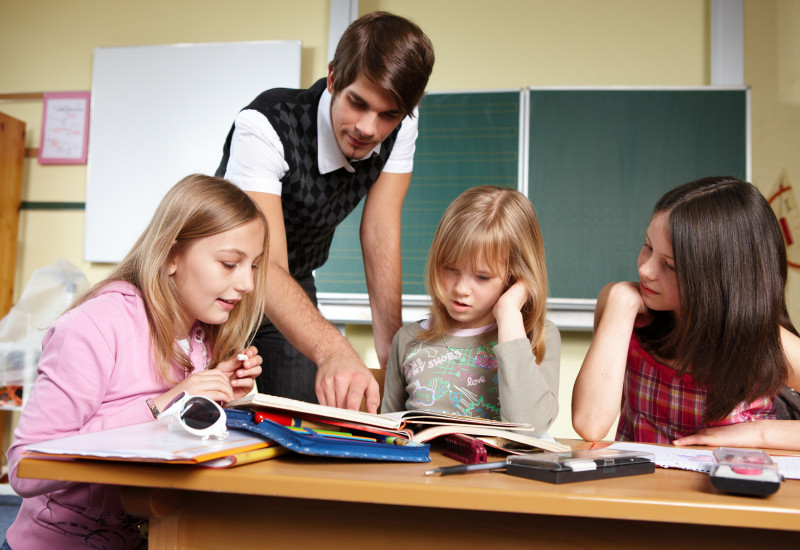 ein junger Leerer und drei Schulkinder sind an einem Tisch in einem Klassenzimmer über Hefte gebeugt