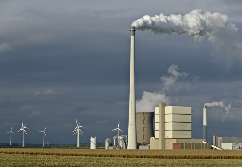 Kraftwerk mit rauchenden Schornsteinen und Windkraftanlagen vor dunkelgrauem Himmel auf einem Feld