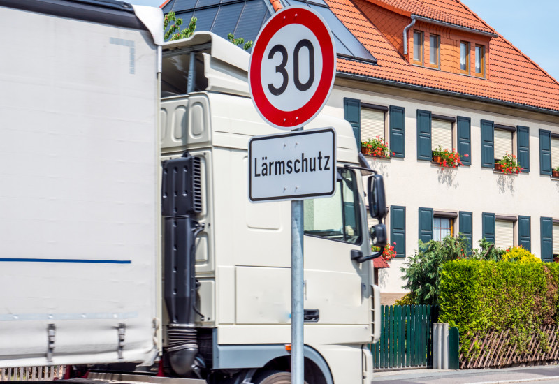 Ein Lkw fährt durch ein Wohngebiet, an der Straße steht ein Verkehrsschild "Tempo 30 Lärmschutz".