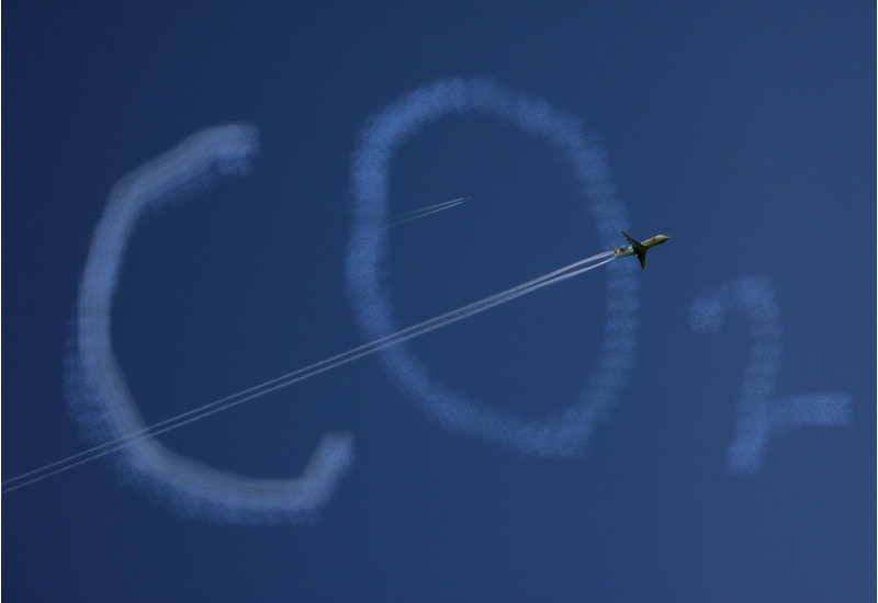an einem blauen Himmel bilden Wolken den Schriftzug "CO2", ein Flugzeug fliegt durch das Bild und zieht einen Kondensstreifen hinter sich her