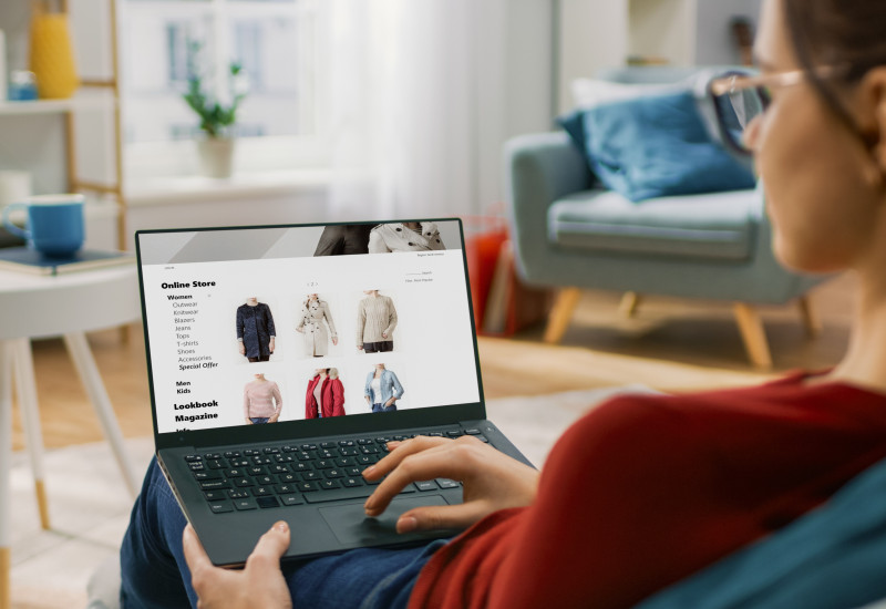 eine Frau sitzt in einem Wohnzimmer auf dem Sofa und schaut auf einem Laptop in einem Onlineshop nach Kleidung