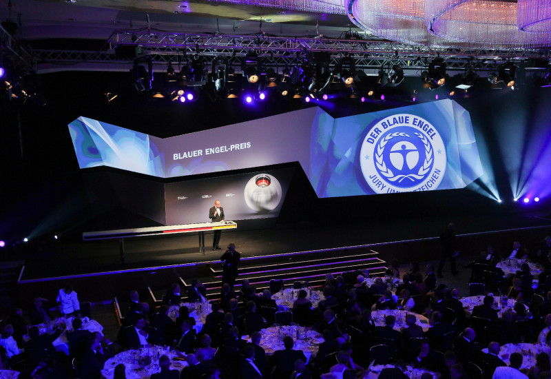 in einem Saal steht ein Sprecher auf der Bühne, im Hintergrund ist das Logo "Blauer Engel" und der Schriftzug "Blauer Engel-Preis" zu sehen