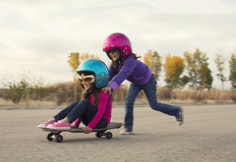 zwei kleine Mädchen mit Sturzhelmen schieben sich gegenseitig auf einem Skateboard auf einem gepflasterten Platz