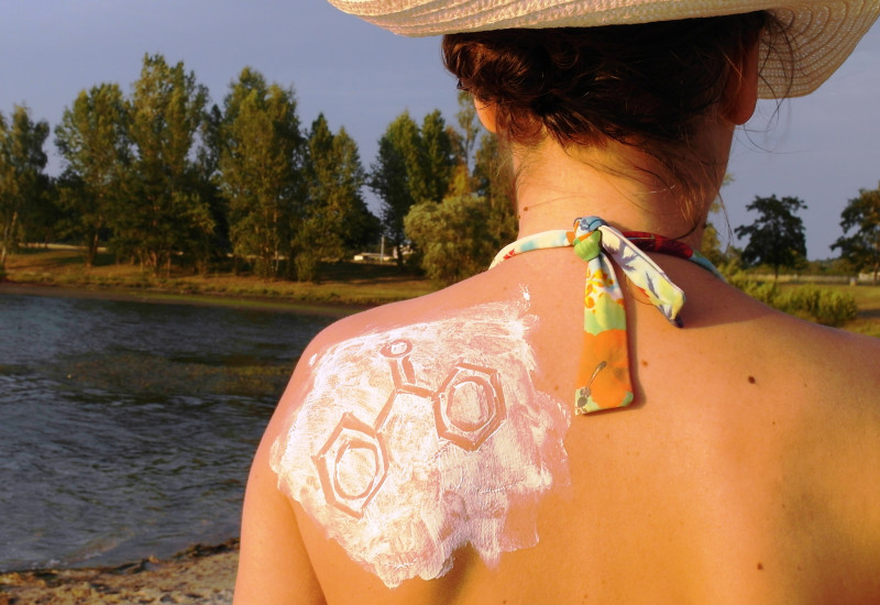 Eine Frau mit Sonnenhut und Bikini steht an einem See. Man sieht Rücken und Hinterkopf. Das linke Schulterblatt ist mit Sonnencreme eingecremt, in die eine chemische Strukturformel gezeichnet ist.