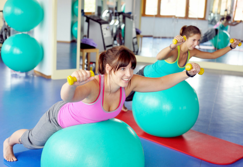 zwei Frauen in einem Sportraum trainieren mit Gymnastikbällen und Hanteln auf einer Matte