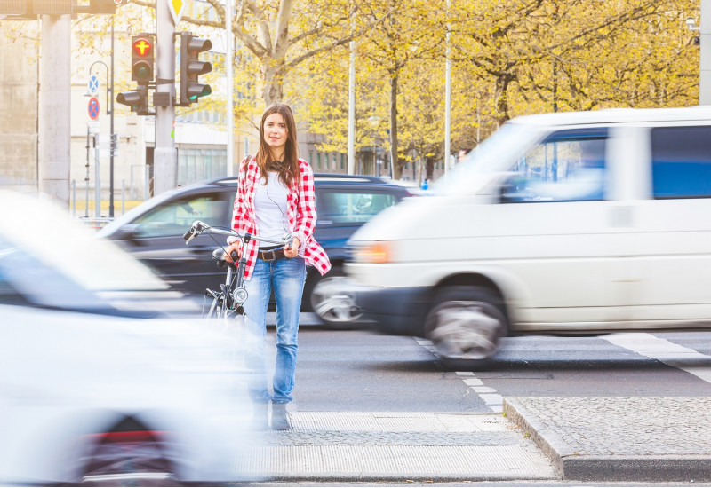 eine junge Frau wartet, ein Fahrrad schiebend, auf einer Mittelinsel an einer roten Ampel, vor und hinter ihr fahren Autos vorbei