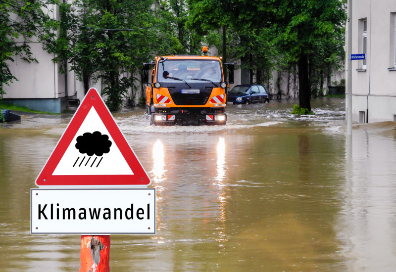 überflutete Straße in der Stadt, davor ein Warnschild "Regen / Klimawandel", ein kommunales Einsatzfahrzeug bahnt sich den Weg durchs Wasser