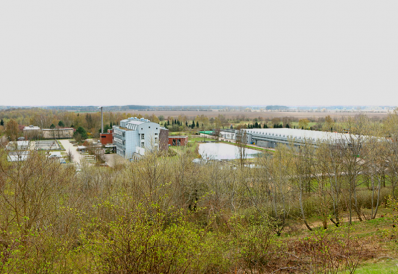 Foto: In Mitten von Bäumen und Feldern ist ein Ensemble aus Gebäuden, einer Halle und Wasserbecken zu sehen.