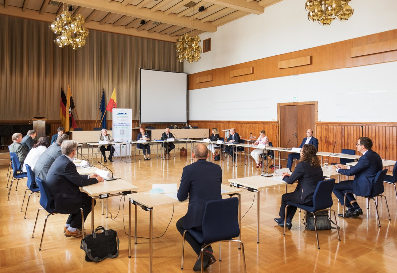 in einem Saal sitzen Männer und Frauen an Tischen im Kreis, hinten ein Aufsteller und Fahnen
