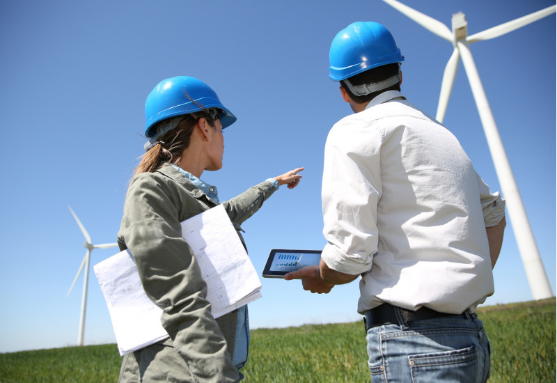 eine Frau und ein Mann mit blauen Bau-Schutzhelmen stehen mit einem Tablet und Papierunterlagen in der Hand auf einer Wiese und zeigen auf eine Windkraftanlage