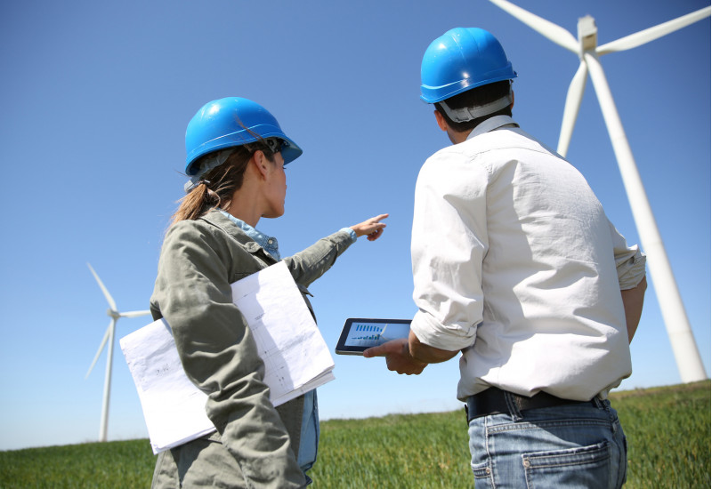 eine Frau und ein Mann mit Bauhelmen, Planunterlagen und einem Tablet schauen sich einen Windpark an