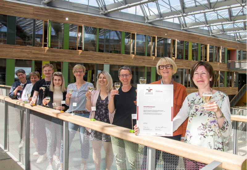 acht Frauen und ein Mann haben sich im UBA Dessau für ein Foto aufgestellt und halten lächelnd Gläser zum Anstoßen und das Zertifikat des Audits Beruf und Familie in die Kamera