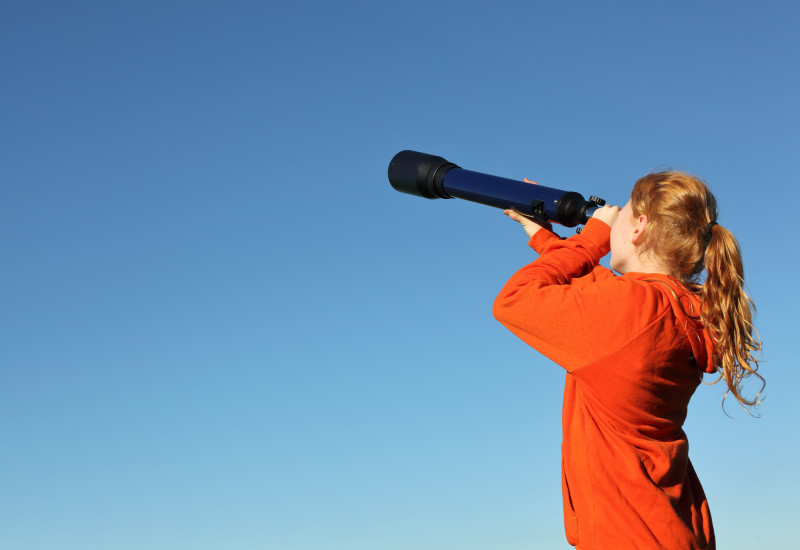 eine junge Frau schaut mit einem Fernrohr in die Ferne, im Hintergrund klarer, blauer Himmel