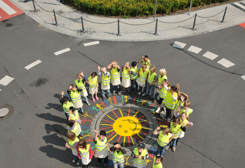 Kinder stehen jubelnd um einen Gullideckel, den sie bunt mit einer Sonne bemalt haben