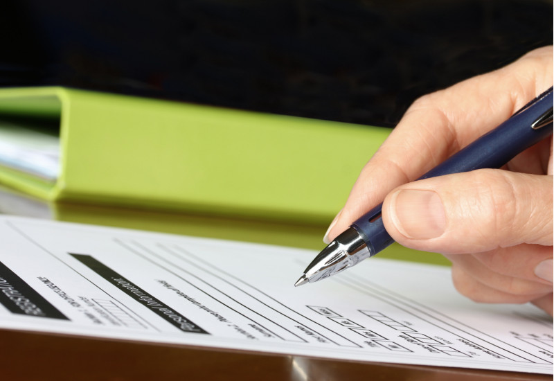 Im Vordergrund Hand hält Stift vor einem Blatt Papier. Im Hintergrund ein grüner Ordner. 
