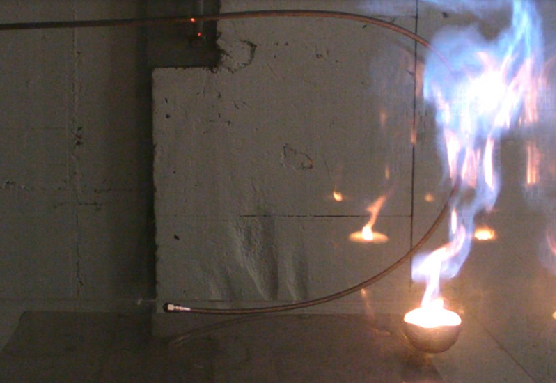 Ein Metallrohr ist U-förmig vor einer Wand gebogen. Auf der rechten Seite lodert eine große Flamme das ganze Rohr entlang.