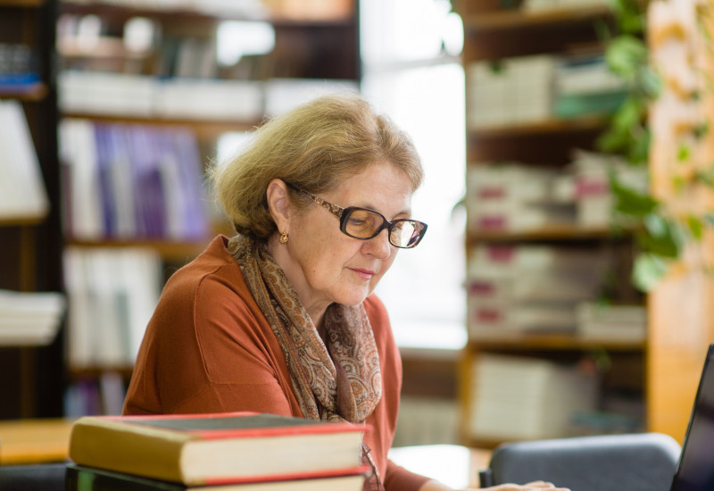 Eine Frau sitzt mit einem Laptop in einer Bibliothek und liest ein Buch.