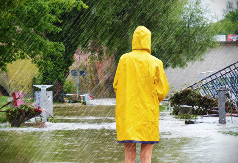 Eine Frau mit Regenmantel steht im Regen vor einer überfluteten Landschaft.