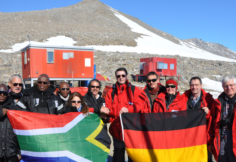 Vor einer Forschungsstation steht eine Gruppe von Männern und Frauen. Links die Gruppe aus Südafrika, recht die deutsche Gruppe. Beide halten ihre jeweilige Landesflagge in den Händen.