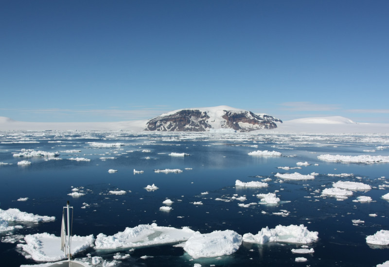 Die Aufnahme ist vom Bug eines Schiffes gemacht, so sind noch der vorderste Teil des Schiffes und dann das Südpolarmeer im Bild. Auf dem Wasser treiben Eisschollen und im Hintergrund ist antarktisches Festland. 