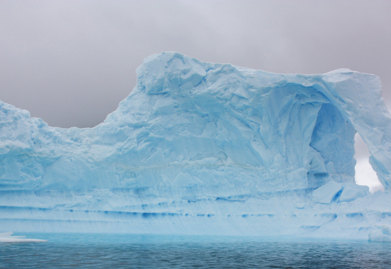 An appealing light blue iceberg floats in the nearly black water. The sky is gray and drab.