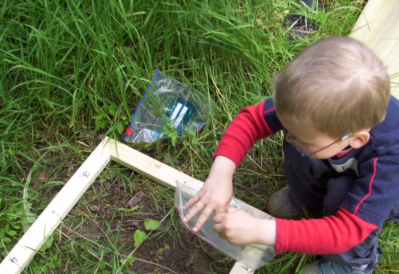 Ein Kind guckt sich den Boden innerhalb eines Holzrahmens ganz genau an und füllt eine Probe in eine Plastiktüte