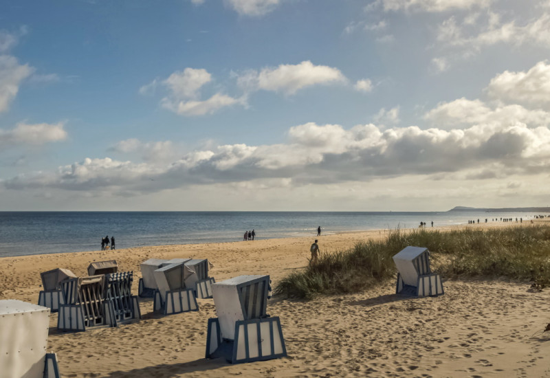 Das Bild zeigt einen Sandstrand an der Ostsee mit Dünen und Strandkörben. Die der oder dem Betrachtenden zugewandten Strandkörbe sind versperrt. Am Horizont ist das Meer zu sehen. An der Wasserlinie sind einzelne Personen und Personengruppen zu erkennen, sonst ist der Stand leer. Der Himmel ist in Teilen bewölkt.