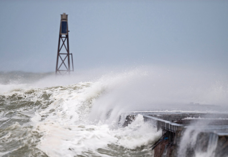 The picture shows a storm surge wave breaking over a coastal building. A metal mast is visible in the background.