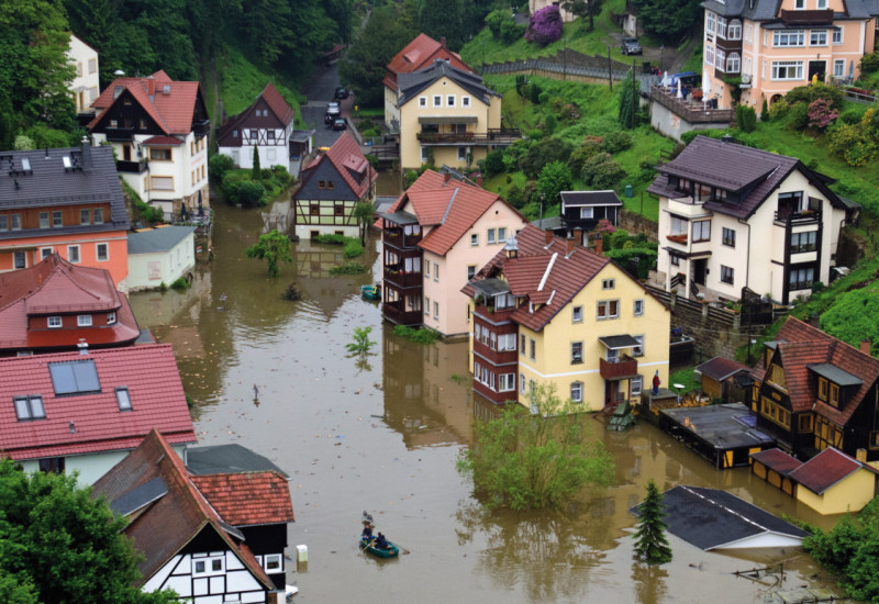 The picture shows a village – situated on the slope of a hill – inundated by floodwater. Some of the lower buildings and some lower floors of houses have been flooded.