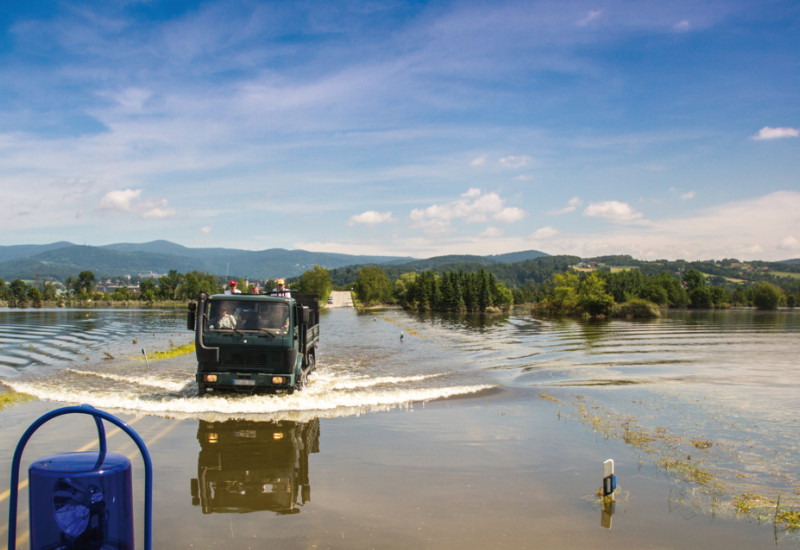 Das Bild zeigt eine von Hochwasser überschwemmte Landschaft. Im Bildzentrum ist eine überschwemmte Straße zu sehen, auf der ein Lastwagen mit Personen auf der Ladefläche fährt. Im Vordergrund des Bildes ist ein Teil eines blauen Einsatzfahrzeugs inklusive blauer Rundumleuchten zu sehen, das ebenfalls auf der überschwemmten Straße unterwegs ist.