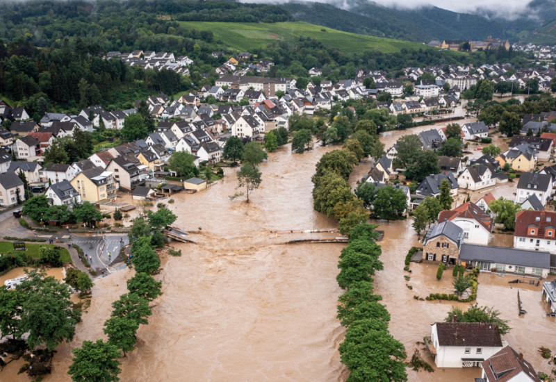 The picture shows a village consisting of single- and multi-family houses in a valley. The river flowing through the village has burst its banks and its brown water has inundated the low-lying parts of the village. The usual river course can be recognised by means of tree lines. In addition, you can see a destroyed bridge which used to cross the river.