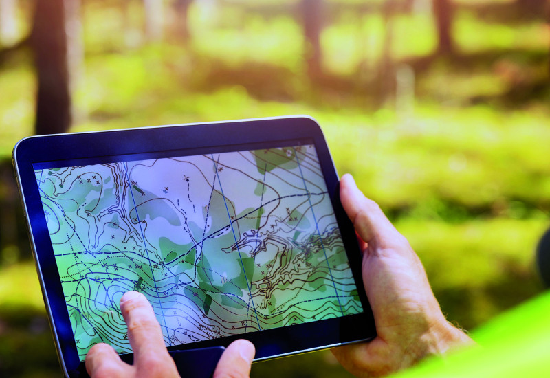 Das Bild zeigt Hände, die ein Tablet halten, auf dessen Bildschirm eine Karte mit Höhenlinien und weiteren Markierungen zu sehen ist. Der verschwommene Bildhintergrund zeigt, dass die Person, die das Tablet hält, in einem Wald steht.