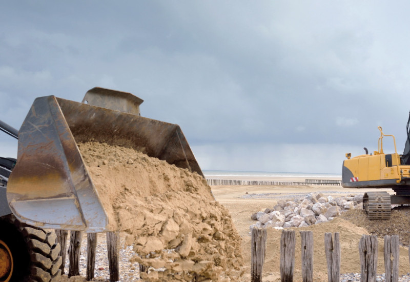 Das Bild zeigt im Vordergrund die gefüllte Schaufel eines Radladers, aus der Sand auf einen Sandstrand entleert wird. Im Hintergrund sind hölzerne Pfähle und eine Steinhalde auf einem weiten Sandstrand zu sehen. Der Himmel ist von dunkelgrauen Wolken verhangen.