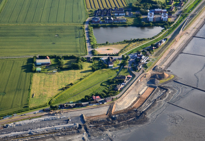 The picture shows an aerial view of a land protection dyke under construction. In the background, there are agricultural areas and houses as well as standing water visible.