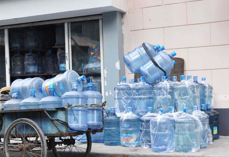 Altes Lastendreirad, welches auf der Straße abgestellt wurde und mit leeren Wasserkanistern vollbepackt ist; auf dem Fußgängerweg lagern weitere leere Plastikkanister; einige sind in Plastiktüten eingepackt, andere nicht; die Kanister kommen aus einem Lager, welches offen ist