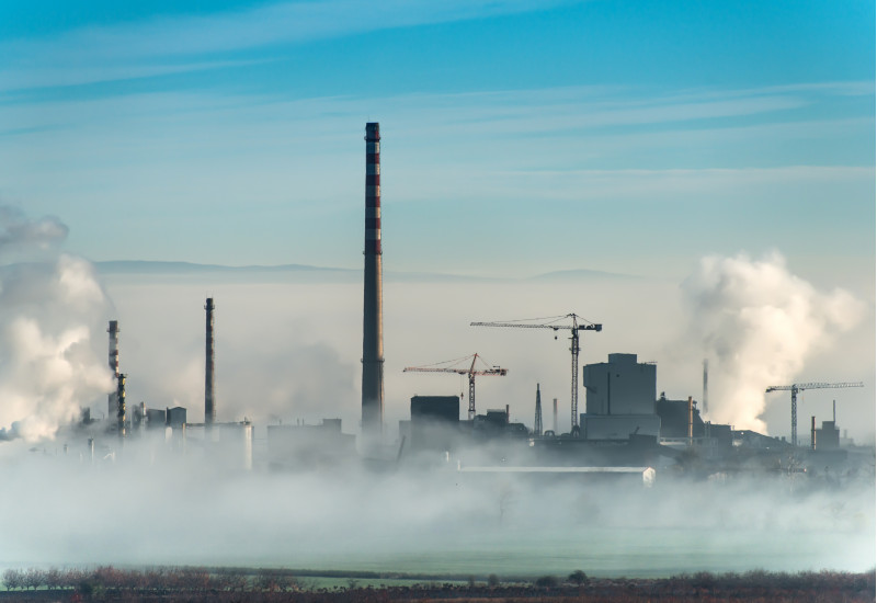 Industrieanlage mit mehreren Rauchtürmen und zwei Baukränen auf dem Gebiet; im Vordergrund steigt Nebel aus dem Kühlwasserflussauf
