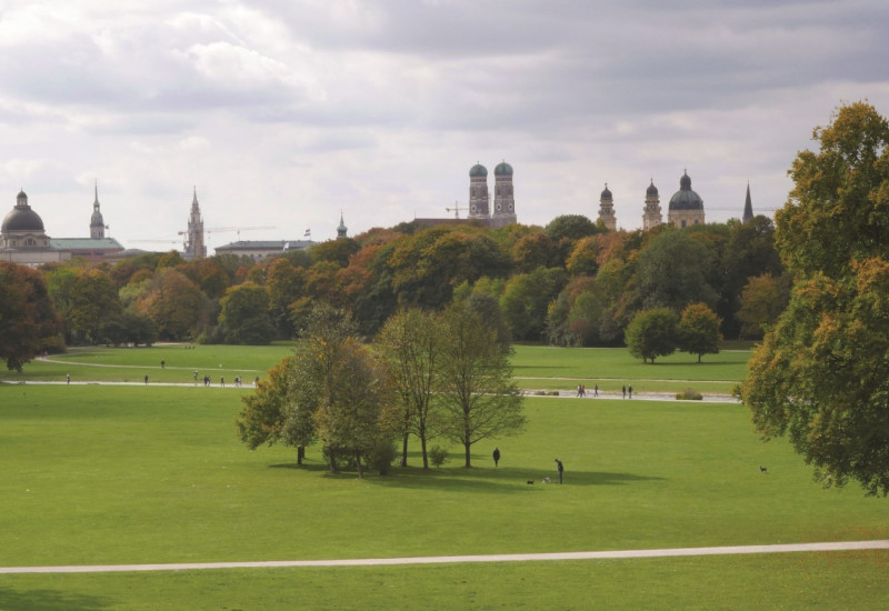 The picture shows a view of the English Garden in Munich. You can see large meadows criss-crossed by paths with individual groups of trees. In the background you can see the silhouette of Munich. 