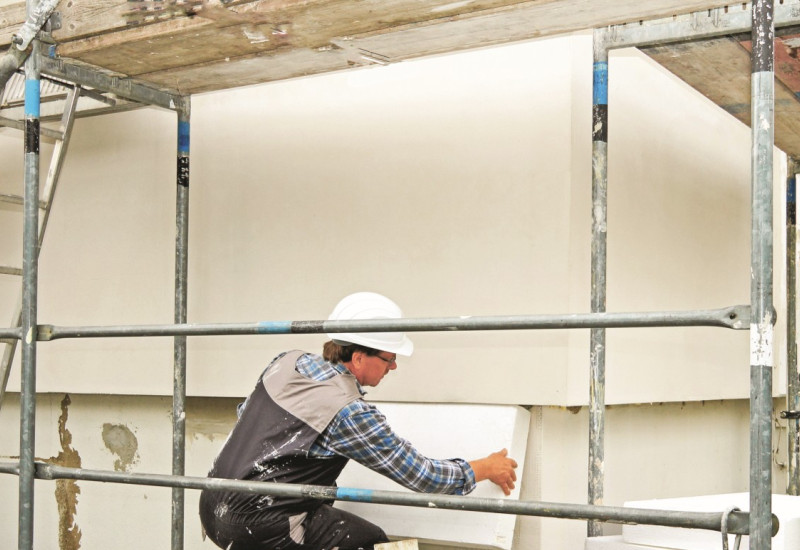 The picture shows a man in a construction helmet squatting on scaffolding and attaching an insulation board to the wall of a house. 