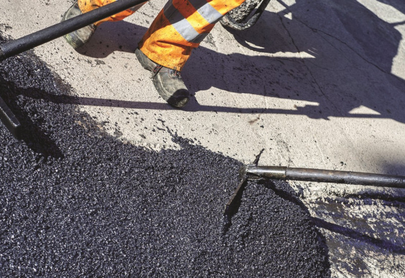 The picture shows the legs of a road construction worker who is repairing a piece of road with fresh asphalt.