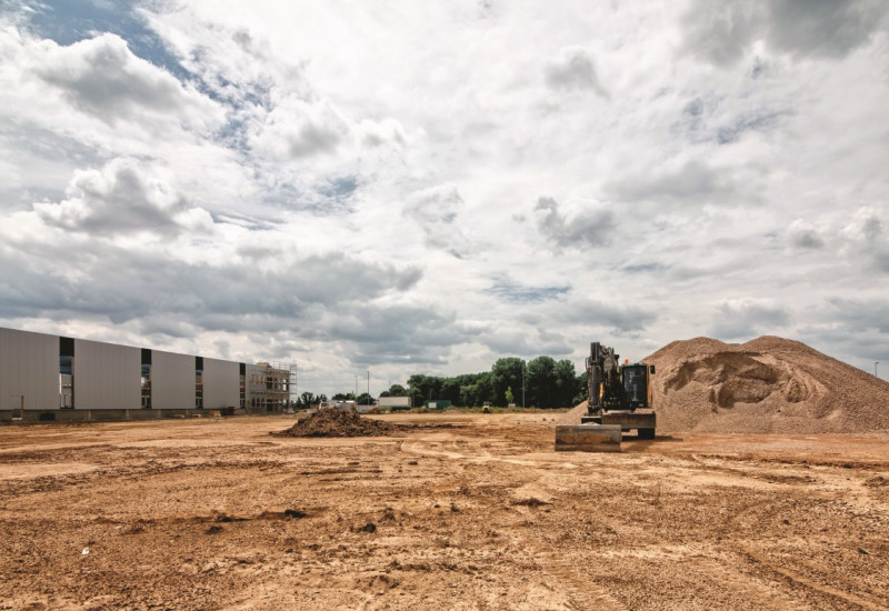 Das Bild zeigt eine große offene Bodenfläche auf einer Baustelle. Im Hintergrund steht rechts neben einem aufgeschütteten Erdhügel ein Bagger auf der Fläche, links sieht man Teile einer Industriehalle. 