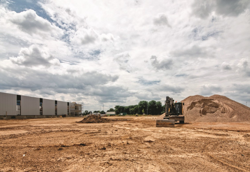 The picture shows a large open ground area on a construction site. In the background, an excavator is standing on the area to the right of a mound of earth, and parts of an industrial hall can be seen to the left. 