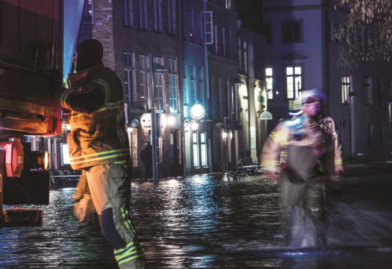 Das Bild zeigt einen mit Wasser überfluteten Straßenzug bei Nacht. Zwei Einsatzkräfte waten durch das Wasser, einer davon befindet sich direkt an einem Einsatzfahrzeug. 