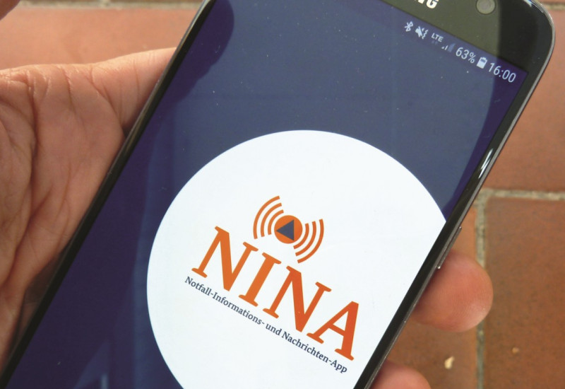 The picture shows a hand holding a smartphone. The NINA app is called up in it. 