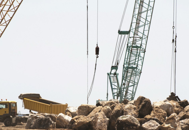 Das Bild zeigt eine große Baumaßnahme am Meer. Von großen Krananlagen und einem großen LKW werden große Steinblöcke bewegt. 