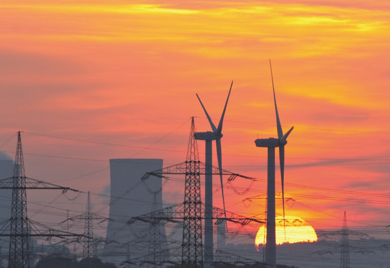 The picture shows two wind turbines, a cooling tower and several electricity pylons against the setting sun.